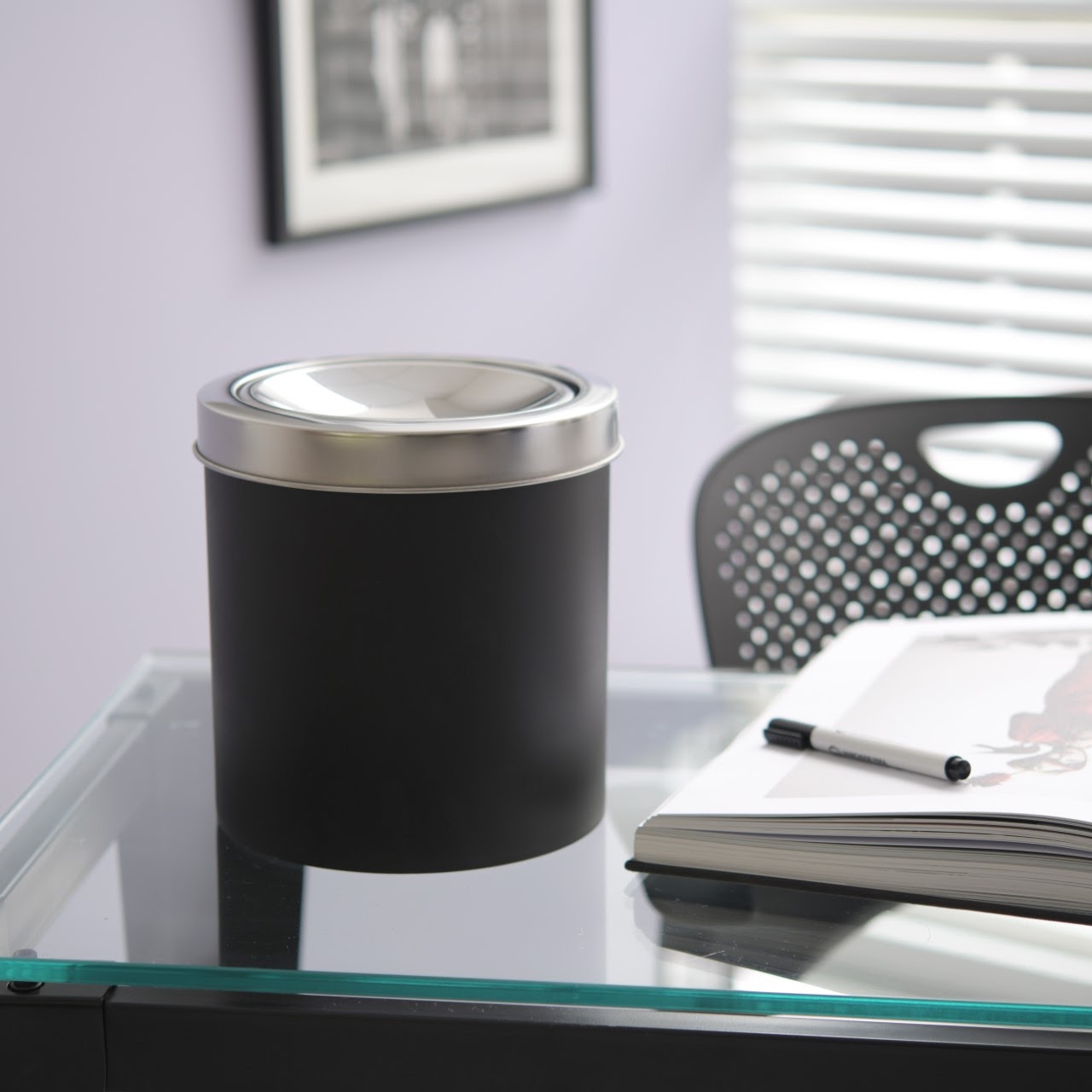 Lixeira basculante Decorline na cor preto. Ela está em cima de uma mesa de vidro de um escritório. 
